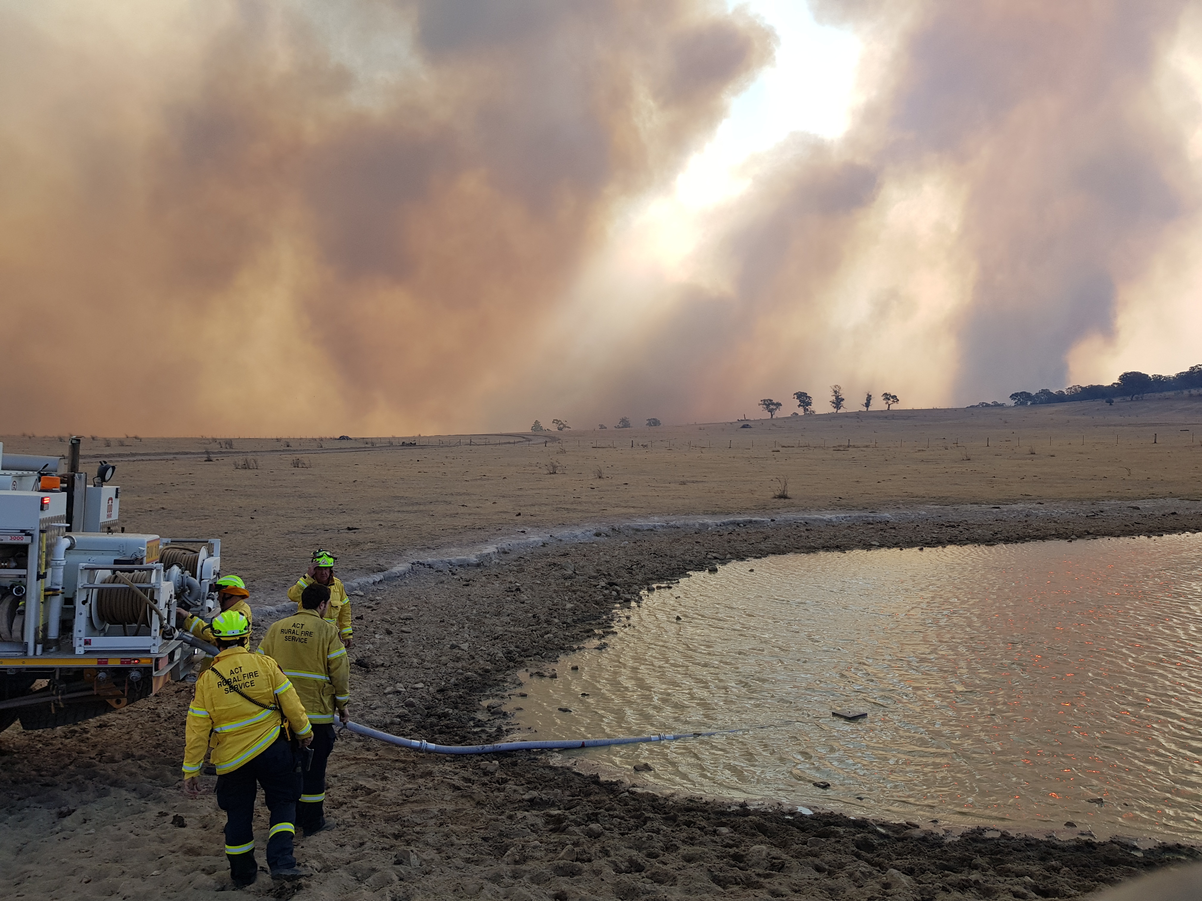 Photograph of firefighters filling their tanker from a dam as smoke fills the horizon.